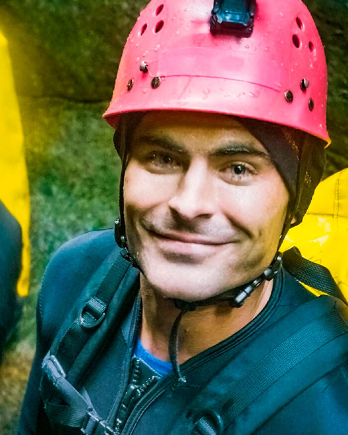 Zac Efron wearing a red helmet and outdoor gear smiling during an adventurous activity in a natural setting.