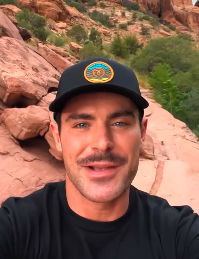 Zac Efron outdoors wearing a black cap, with a natural rocky background, showing his face in a casual selfie.