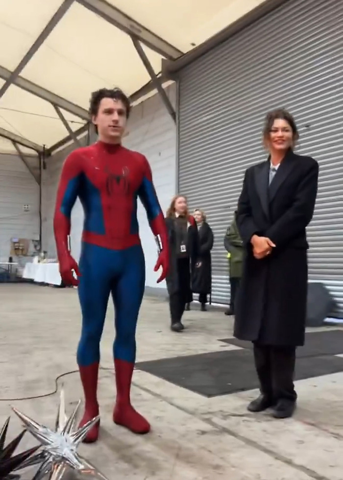 Tom Holland in Spider-Man suit standing beside Zendaya in black coat on set, sparking leaked video pregnancy rumors.