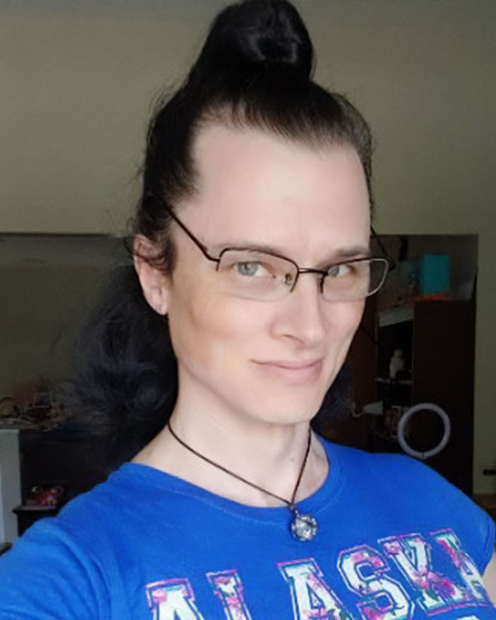High school teacher smiling indoors wearing glasses and a blue Alaska shirt with long dark hair tied up.