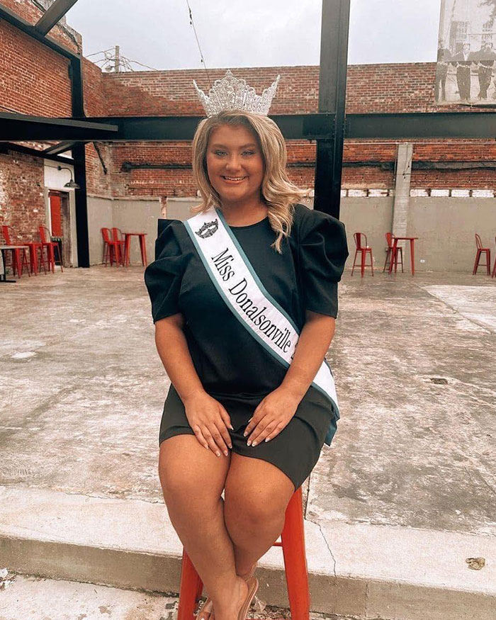 Beauty queen wearing crown and sash sitting on a red stool in an industrial outdoor setting, facing the camera.