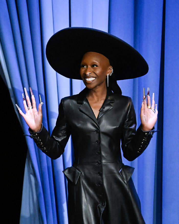 Cynthia Erivo wearing a black leather outfit and large hat, showing her oddly large hands during a public appearance.