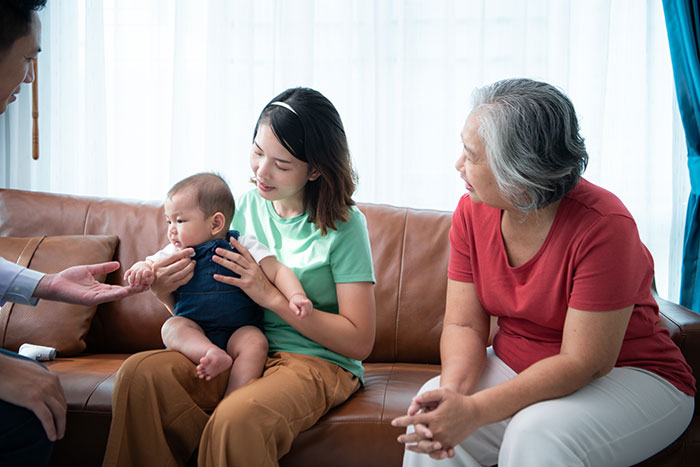 Mother-in-law expressing concern to daughter-in-law holding premature baby while sitting with family on a couch. Mother-in-law expressing concern to daughter-in-law holding premature baby while sitting with family on a couch.