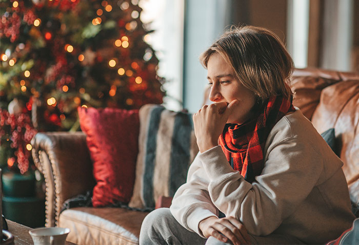 Young woman sitting on a couch looking thoughtful during a Christmas gathering in a cozy living room decorated for the holiday season.