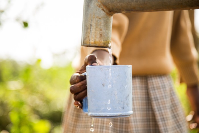 Person in African country filling a metal cup with water from a pipe, embracing a simple lifestyle outdoors.
