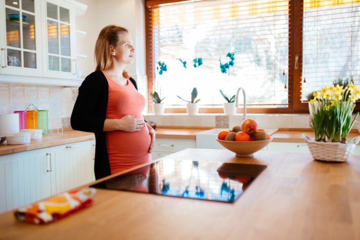 Pregnant woman standing in a bright kitchen, reflecting on family issues as husband won&rsquo;t stand up to his mother.
