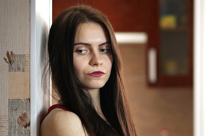 Teen girl with long dark hair looking thoughtful and concerned, wondering about missing $30k, indoors near a wall.