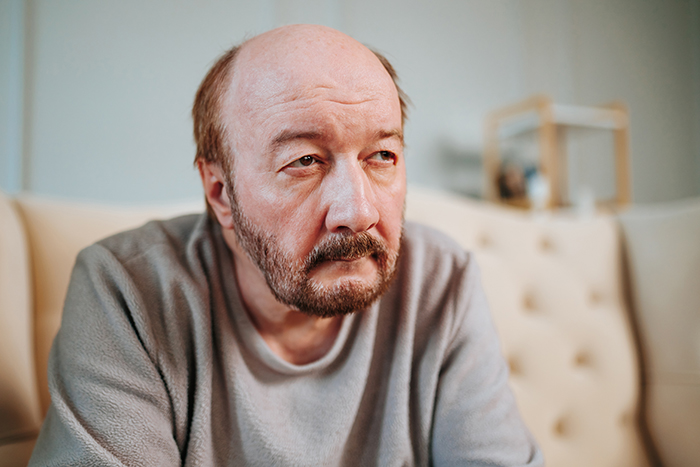 Older man with a serious expression sitting on a couch, reflecting themes of marriage issues and banned family members.