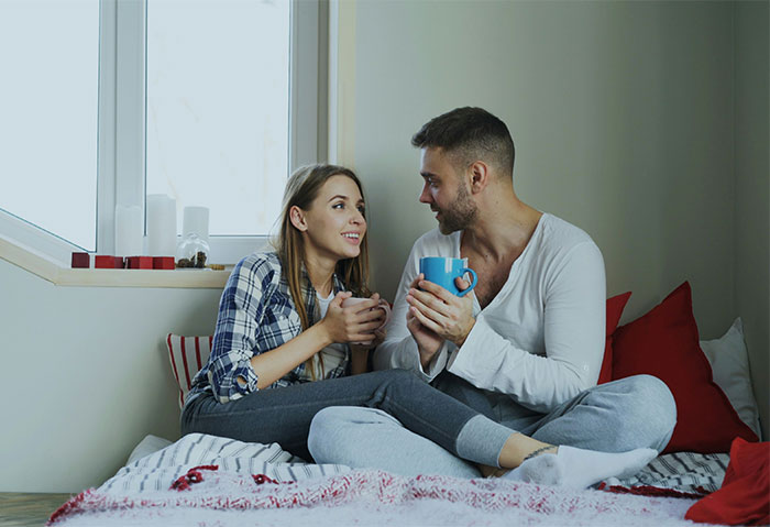 Couple sitting on bed drinking coffee, woman looks happy while man appears to be explaining, relationship built on lies concept.