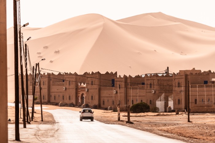 Car driving toward traditional buildings near large sand dunes in an African country embracing a simple lifestyle.