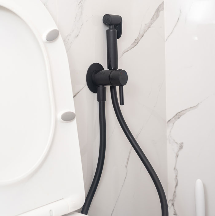 Black handheld bidet sprayer faucet mounted on marble wall beside white toilet seat in bathroom setting.