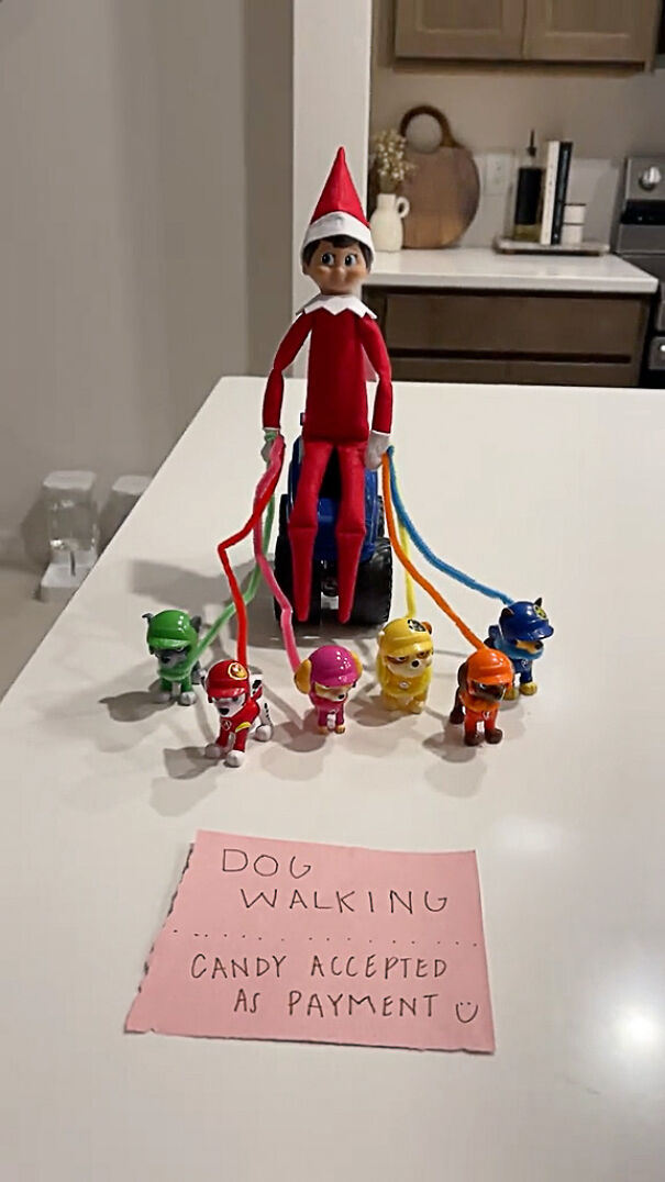 Elf on the Shelf holding toy dogs on leashes with a note about dog walking and candy payment on a kitchen counter.