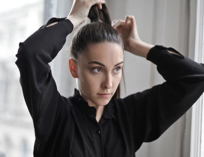 Young woman tying her hair back near a window, showing a focused expression related to spicy roasts by little kids.