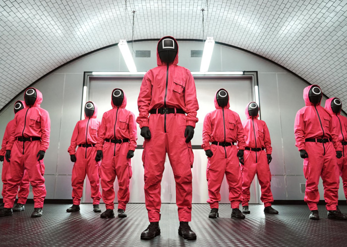 Group of people in red costumes and black masks resembling Squid Game characters in a dimly lit room setting.
