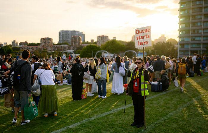 Crowd gathered on grassy public space as a staff member holds a sign marking the start of a queue on New Year’s Eve. Crowd gathered on grassy public space as a staff member holds a sign marking the start of a queue on New Year’s Eve.