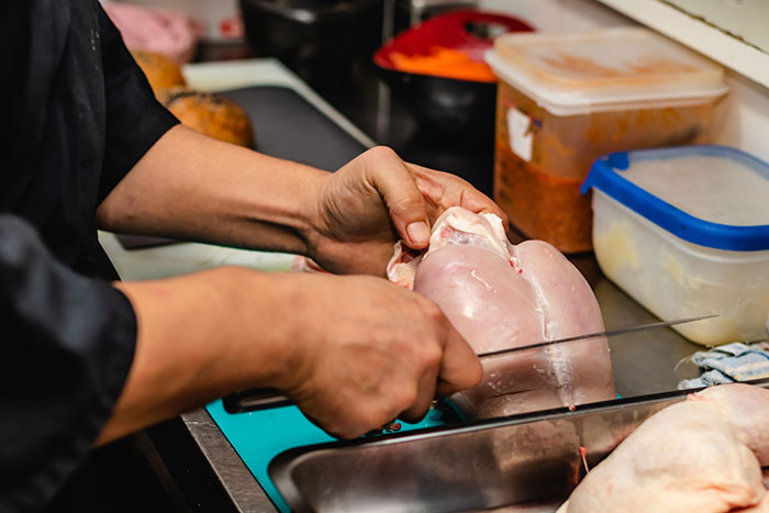 Hands slicing raw chicken breast on a cutting board in a kitchen, highlighting concerns about how chicken is prepared.