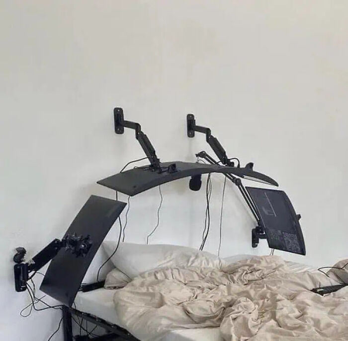 Multiple black monitors with flexible arms floating above an unmade bed, creating a trippy pictures illusion.