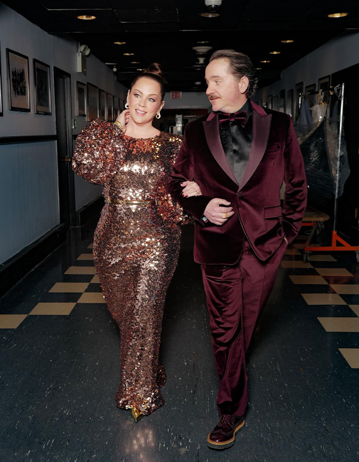 Melissa McCarthy wearing a sparkling gown walking arm-in-arm with a man in a velvet suit, showcasing her weight loss transformation.