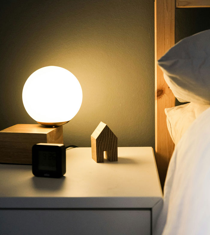 Bedside table with a glowing round lamp, a small wooden house ornament, and a digital clock at night.