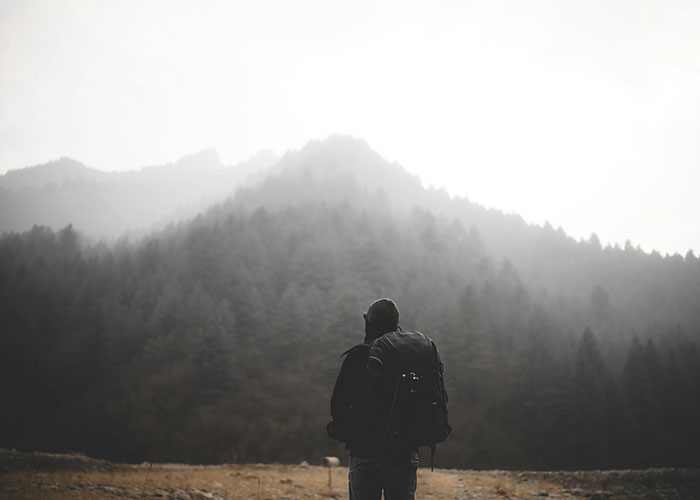 Person with backpack standing in foggy remote forested mountains, evoking creepy and mysterious isolated place vibes.