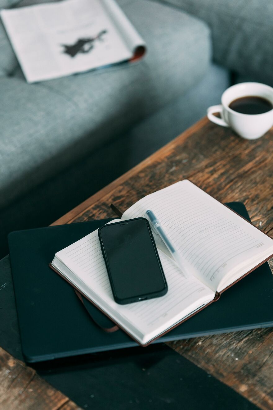 Smartphone on an open notebook with a pen, laptop below, and coffee cup on a wooden table for good morning message. Smartphone on an open notebook with a pen, laptop below, and coffee cup on a wooden table for good morning message.