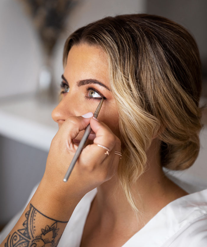 Woman having eye makeup applied with brush by tattooed hand, highlighting things women do to seem attractive men dislike.