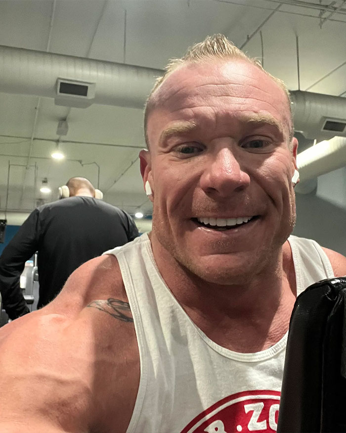 Muscular man smiling in a gym, wearing a white tank top and earbuds, with focus on his face and shoulders. Muscular man smiling in a gym, wearing a white tank top and earbuds, with focus on his face and shoulders.