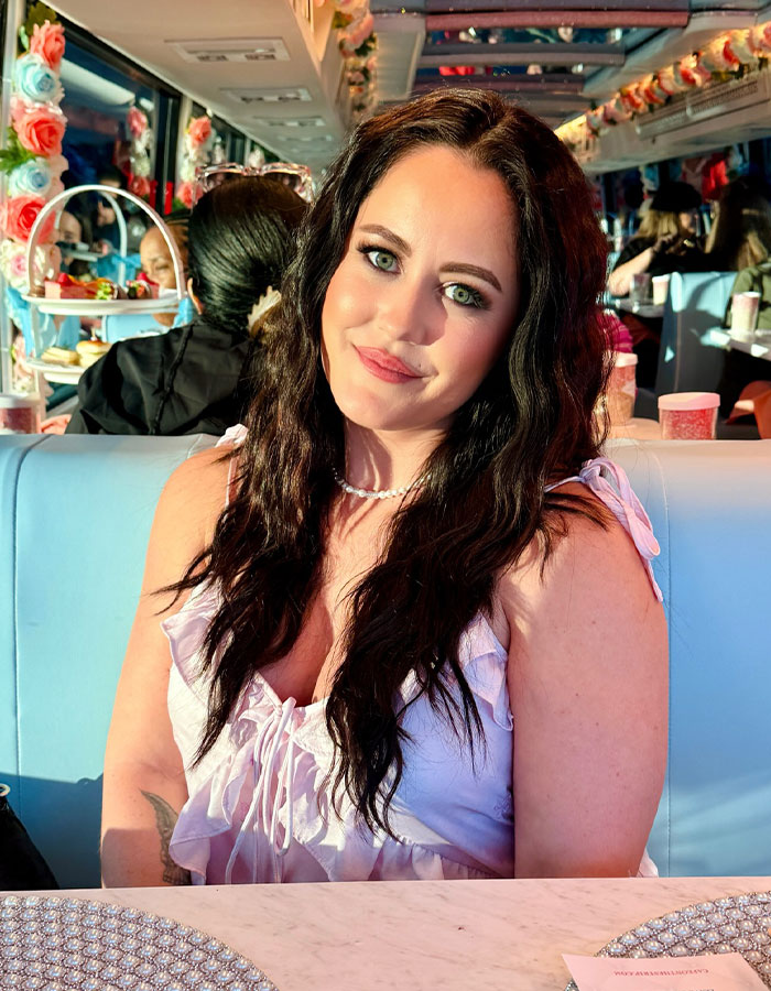 Jenelle Evans with long dark hair, wearing a lavender top, smiling while seated in a colorful restaurant setting. Jenelle Evans with long dark hair, wearing a lavender top, smiling while seated in a colorful restaurant setting.