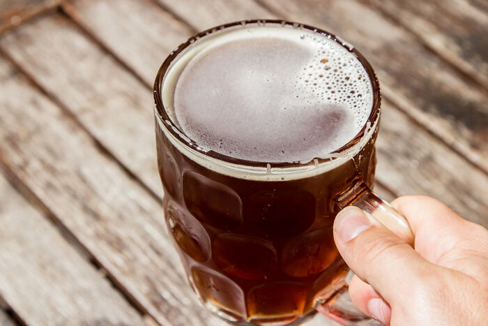 Hand holding a frothy dark beer mug on a wooden table, illustrating revenge stories with a bitter aftertaste theme.
