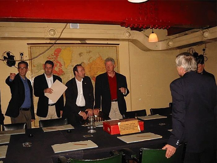 Group of men in a room with a map on the wall and a toddler's foot chillingly visible near Epstein in the photo.
