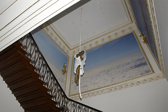 Staircase with ornate railing and ceiling mural showing a hanging sculpture resembling a chilling toddler's foot.