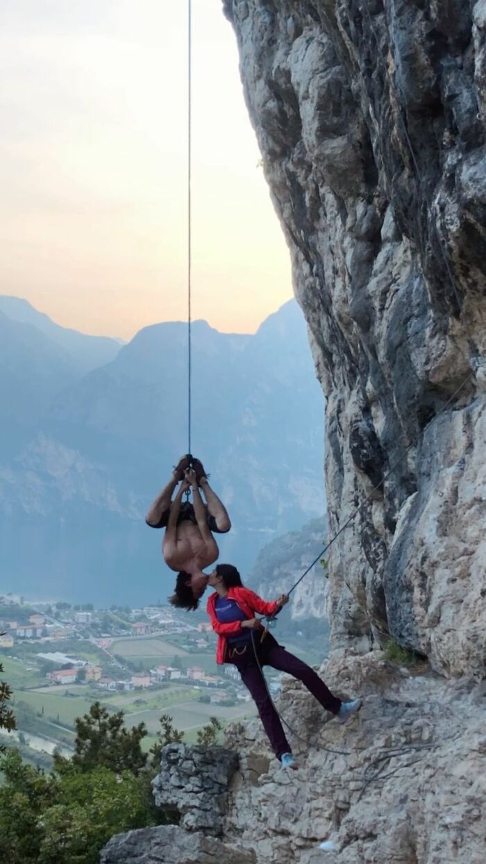Two climbers sharing a moment hanging on a steep rock face with mountains and a valley below in a trippy pictures style.