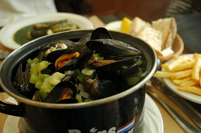 Black pot filled with cooked mussels and chopped vegetables on a table with fries and bread, people share historical facts.