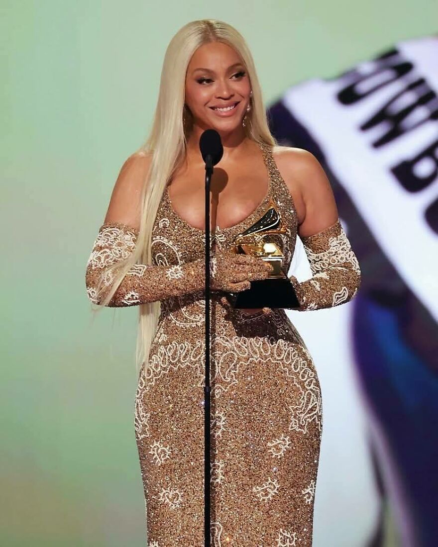 Beyoncé holding a Grammy award on stage, wearing a sparkling gold dress and gloves, celebrating pop culture moments 2025.