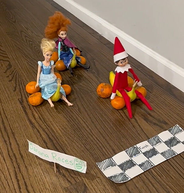 Elf on the Shelf sitting on bananas and oranges as race cars with a handmade finish line on a wooden floor
