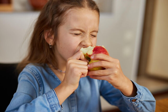 Young girl biting into an apple with eyes closed, capturing a moment of specific and strange kid insult expression.