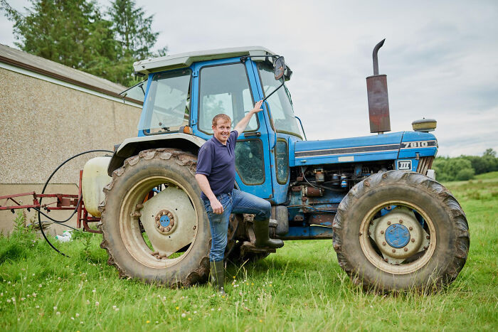 Man standing beside a blue tractor in a grassy field, representing a scene related to family drama situations.