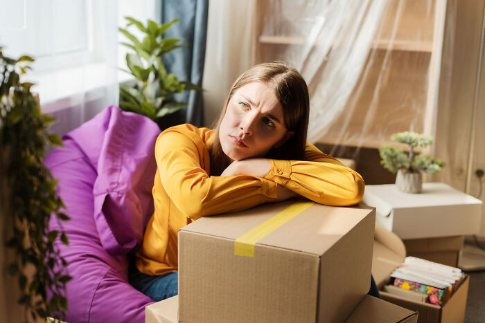Woman in a yellow shirt looking thoughtful, resting on moving boxes amid unintentional big family drama moments.