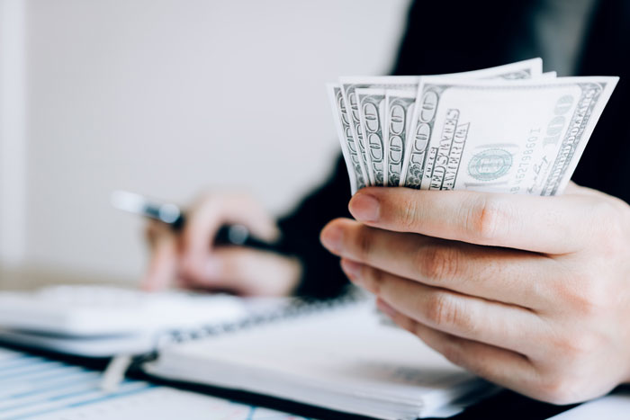 Person holding several US dollar bills while working at a desk, highlighting office scandals impacting workplaces.