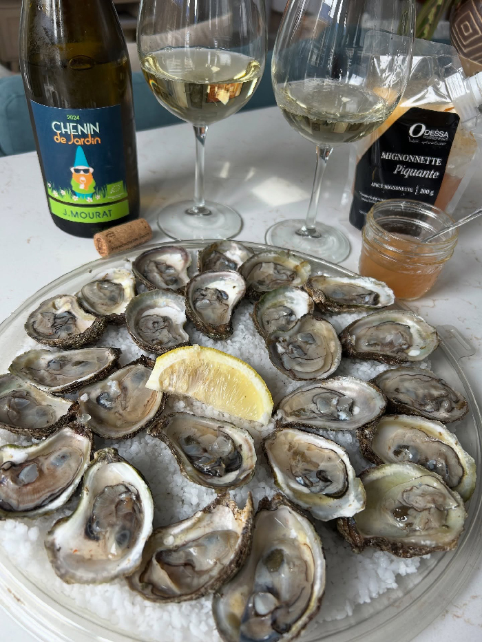 Plate of fresh oysters with lemon wedge, two glasses of white wine, and a bottle on a white marble table in NYC.