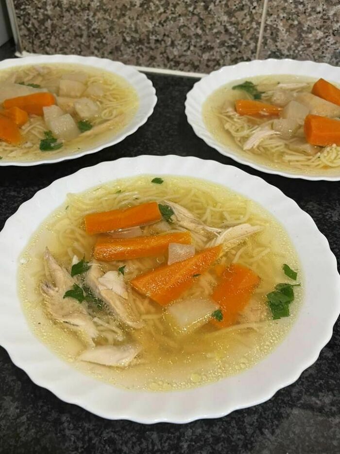 Three bowls of traditional Eastern European chicken soup with noodles, carrots, and herbs on a dark countertop.
