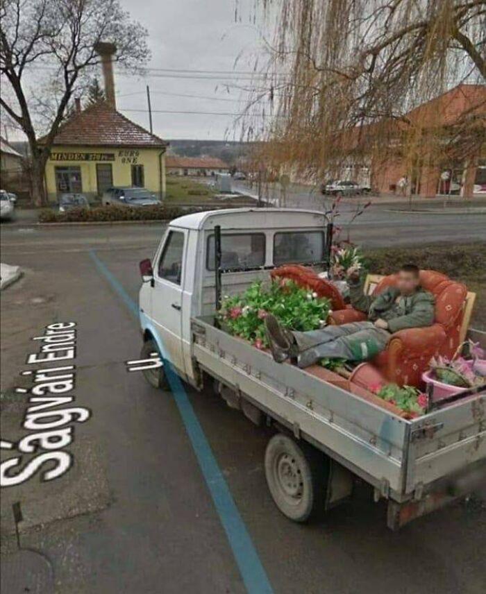 Man lounging on a couch in the back of a small truck filled with plants on a street in Eastern Europe.