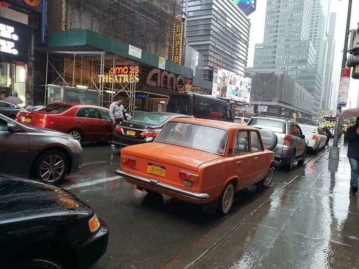 Old orange car parked on a rainy city street with busy traffic, capturing Eastern Europe nostalgia and humor.
