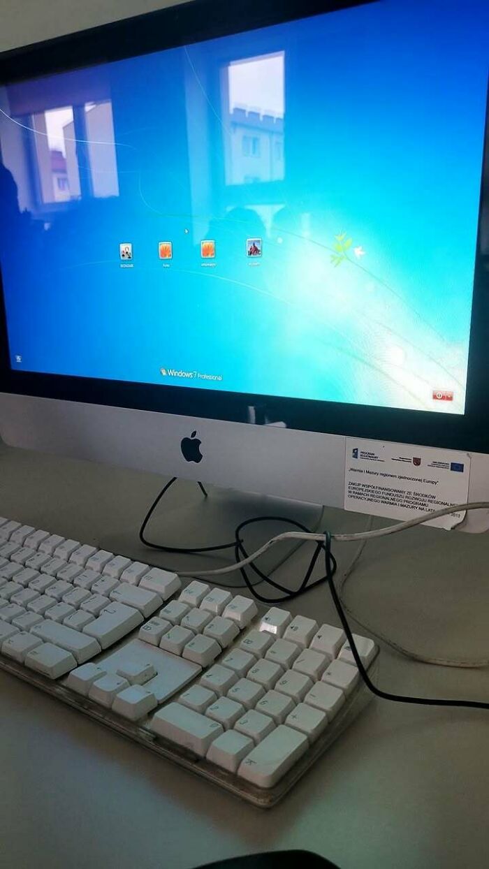 Apple iMac displaying Windows 7 Professional login screen with multiple user profiles and an old keyboard on desk, Eastern Europe vibe.