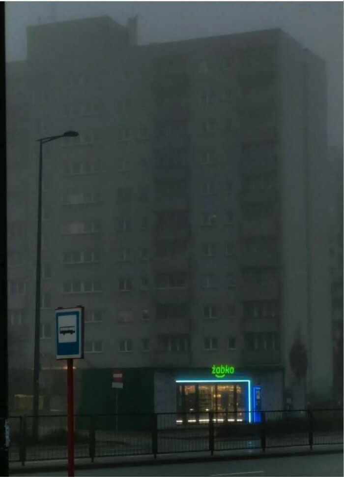 Dimly lit Eastern Europe apartment block with a neon-lit Żabka convenience store in foggy urban setting at dusk