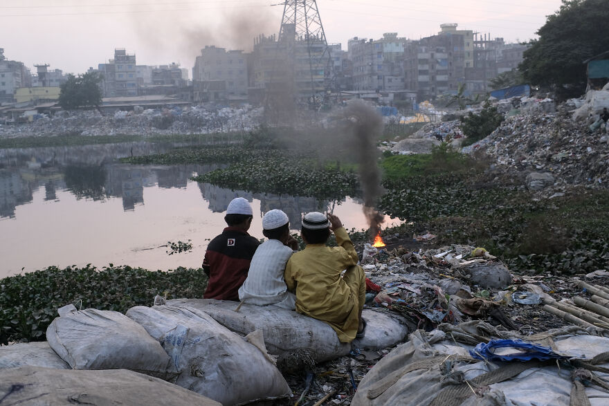 Three boys sitting near polluted water with burning trash and dense smoke, highlighting pollution in top dirtiest countries.