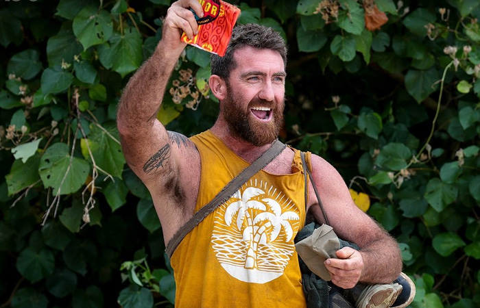 Man in a sleeveless yellow shirt with tattoos raising a red item, celebrating outdoors surrounded by lush green leaves, longest running TV shows.