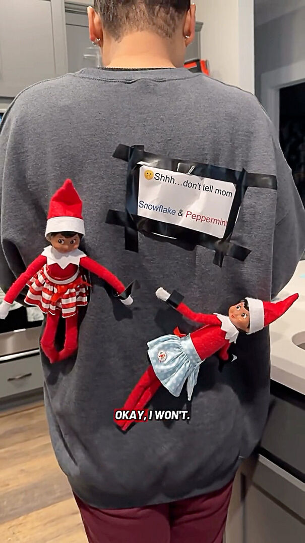 Person wearing a gray sweatshirt with two Elf on the Shelf dolls taped to the back in a kitchen setting.