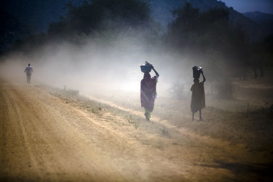 Two people walking on a dusty road carrying containers on their heads with pollution and haze in a rural setting.