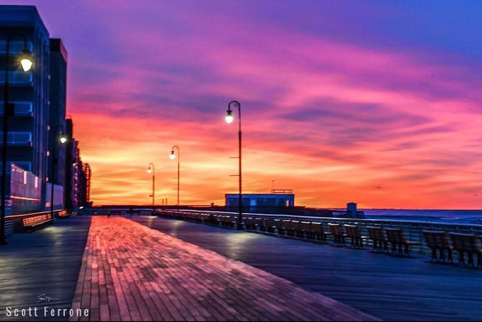Sunset over an empty NYC boardwalk with glowing street lamps and a vibrant purple and orange sky.